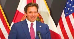 Ron DeSantis is standing at a podium, smiling. Behind him are the flags of the United States and Florida.