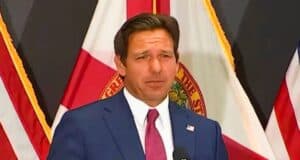 Ron DeSantis in a blue suit and red tie is speaking at a podium with microphones. Behind him are the flags of the United States and Florida. The background includes additional dark-colored flags.
