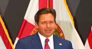 Ron DeSantis is standing in front of a backdrop featuring red and white flags. He has short brown hair and is speaking or making a gesture with his right hand.