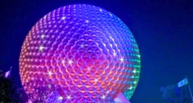 A large geodesic sphere, reminiscent of Remo Saraceni's imaginative designs, is illuminated with a gradient of colors from purple to orange, creating a vibrant display against the dark night sky. The surface is composed of triangular panels with twinkling lights. Flags and trees are partially visible nearby.