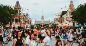 A bustling theme park scene unfolds along Main Street America, with a crowd of people walking past buildings adorned with festive bunting and American flags. Some visitors rest on the ground, while a cloudy sky looms overhead.