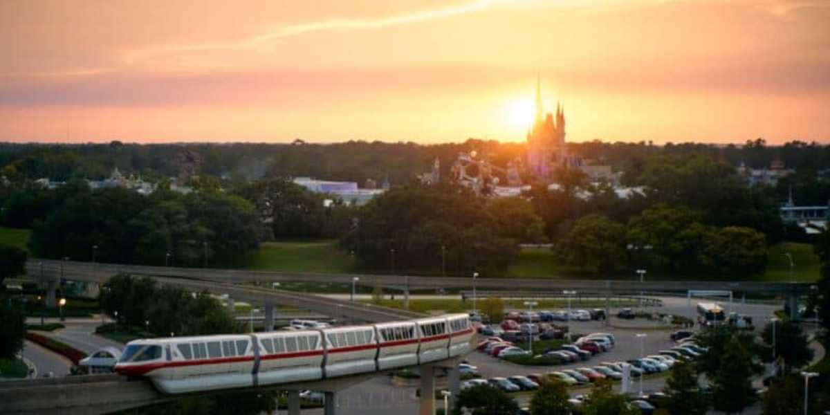 A monorail travels along elevated tracks in the foreground, with a vast parking lot below. In the background, trees surround a castle-like structure, and the sky is illuminated by a vibrant sunset.