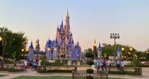 A view of Cinderella Castle at dusk in Walt Disney World Free. The castle is illuminated with blue and gold lights, surrounded by people. Trees and lampposts are visible, with the sky gradually transitioning to night. The scene captures a moment of magic and wonder.