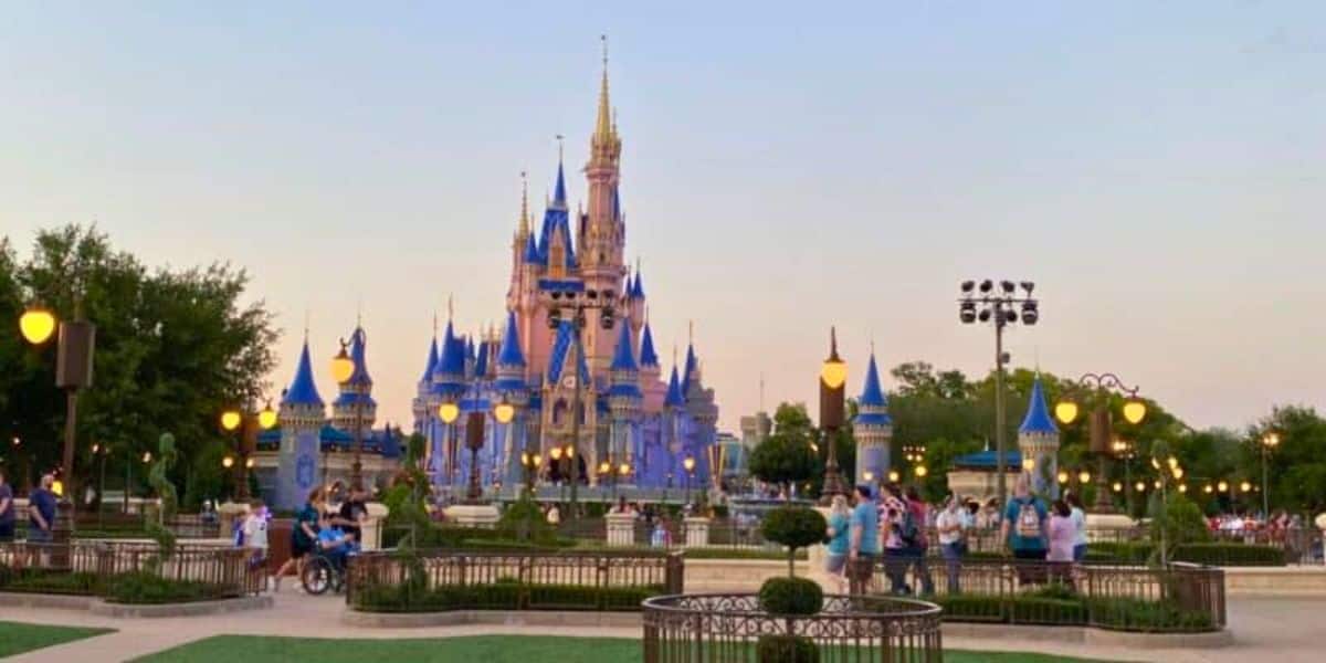 A view of Cinderella Castle at dusk in Walt Disney World Free. The castle is illuminated with blue and gold lights, surrounded by people. Trees and lampposts are visible, with the sky gradually transitioning to night. The scene captures a moment of magic and wonder.