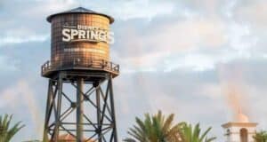 A tall water tower with "Disney Springs" written on it stands against a partly cloudy sky. Palm trees and a white building with a bell tower are visible in the background.