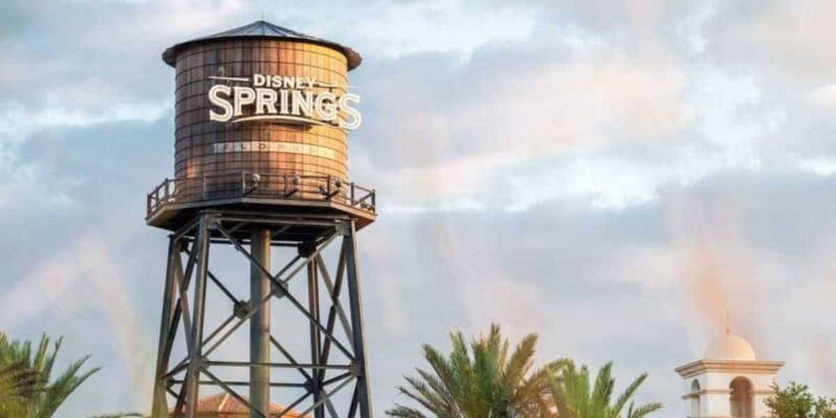 A tall water tower with "Disney Springs" written on it stands against a partly cloudy sky. Palm trees and a white building with a bell tower are visible in the background.