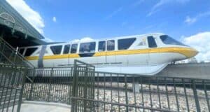 A Disney monorail train in yellow and white colors passing by on an elevated track at a Disney theme park, with a partly cloudy blue sky in the background and a metal fence in the foreground.