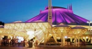 Nighttime view of the Space Mountain attraction at Disneyland, illuminated in purple and white lights. The structure features a futuristic design with a prominent pointed rooftop. Guests can be seen queuing under the brightly lit entrance canopy as Disney prepares to close for the day.