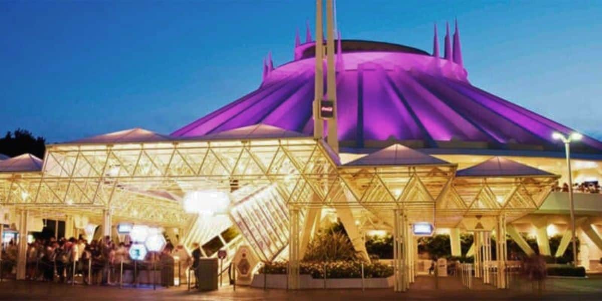 Nighttime view of the Space Mountain attraction at Disneyland, illuminated in purple and white lights. The structure features a futuristic design with a prominent pointed rooftop. Guests can be seen queuing under the brightly lit entrance canopy as Disney prepares to close for the day.
