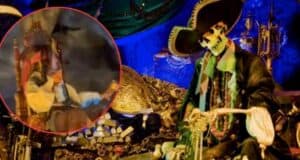 A pirate-themed display featuring a skeleton dressed in pirate attire, seated among treasure chests and gold coins. The background is adorned with blue and green fabrics. There is an inset image of a costumed pirate drinking from a mug.