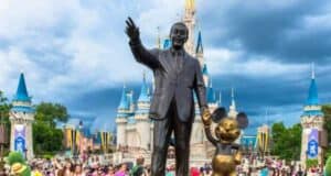 The statue of Walt Disney and Mickey Mouse stands proudly in front of Cinderella's Castle at Disney World, surrounded by Disney guests and vibrant blue skies with scattered clouds.