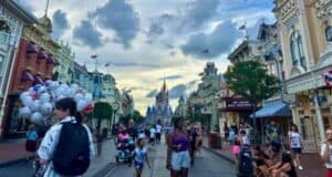 A bustling street scene at a theme park featuring people walking down the main street with the iconic castle in the background. Balloons, decorated shopfronts, and flags add to the festive atmosphere. The sky is partly cloudy, capturing a lively and vibrant moment.