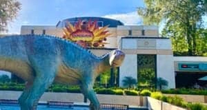 A dinosaur sculpture stands in front of the Dinosaur attraction at Disney's Animal Kingdom. Behind it, a building entrance has a sign that reads "Dinosaur" with a fiery design and a smaller sign that reads "The Dino Institute." As trees and greenery surround the area, whispers about rides closing add to the air of suspense.