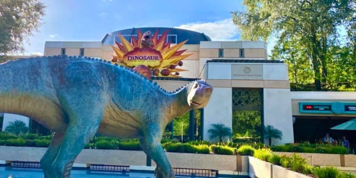 A dinosaur sculpture stands in front of the Dinosaur attraction at Disney's Animal Kingdom. Behind it, a building entrance has a sign that reads "Dinosaur" with a fiery design and a smaller sign that reads "The Dino Institute." As trees and greenery surround the area, whispers about rides closing add to the air of suspense.
