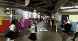 A bustling underground utilidor with a colorful mural and structural pipes overhead. People in a mix of professional and casual attire walk through the space, while a few individuals in uniform stand against the wall. A large "STOP" sign is visible on the floor.