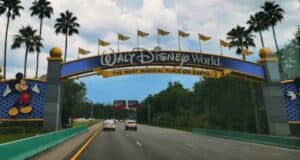 A road leading to the entrance of Walt Disney World is shown, with a large, colorful archway overhead. The arch features the Walt Disney World logo, the phrase "The Most Magical Place on Earth," and images of Mickey Mouse on the left and Minnie Mouse on the right. Tiana's Bayou Adventure awaits inside.