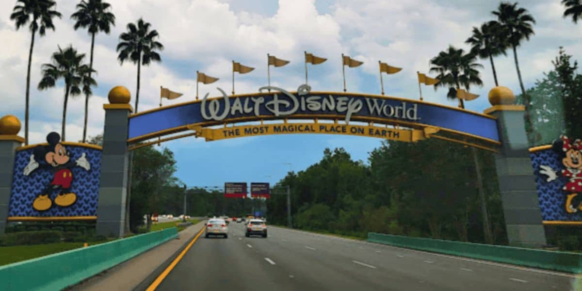 A road leading to the entrance of Walt Disney World is shown, with a large, colorful archway overhead. The arch features the Walt Disney World logo, the phrase "The Most Magical Place on Earth," and images of Mickey Mouse on the left and Minnie Mouse on the right. Tiana's Bayou Adventure awaits inside.
