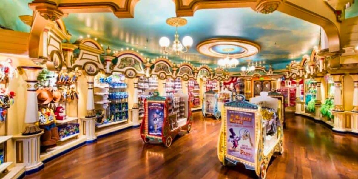 A brightly lit, colorful toy store with wooden floors and ornate architecture. Shelves are lined with a variety of toys, including stuffed animals and action figures. Display stands in the center feature themed Disney merchandise. A painted sky ceiling adds to the whimsical atmosphere.