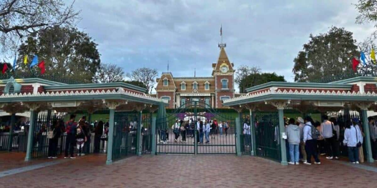 A large crowd of guests in line outside the gates waiting to enter Disneyland Park with the Main Street station of the Disneyland Railroad in the background. Each turnstyle is staffed by a Disney cast member.