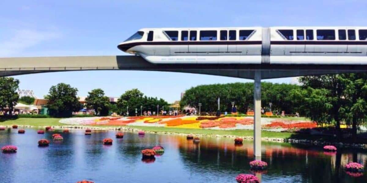 A monorail glides over a scenic park with vibrant flower beds and floating floral decorations on a sunny day.