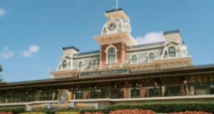 The image captures the entrance to a theme park, reminiscent of Disney magic, with a grand Victorian-style train station and clock tower. Decorative elements stand out against a clear blue sky, while lush landscaping with red flowers adorns the foreground. Nearby, Disney Child Care Services ensures family-friendly fun.