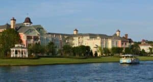 A serene lake scene with a charming, multi-colored building complex in the background, featuring pastel hues reminiscent of Disney Resort pools. A gazebo is on the left near the water's edge. A blue and yellow boat floats on the calm lake to the right. Trees and greenery surround the area.