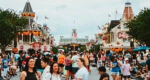 A bustling street scene at a theme park, decorated with orange and yellow autumnal decor. Numerous people are walking, some seated on the ground, while others take photos. Historic-style buildings with flags and signage line the street. The sky is overcast.