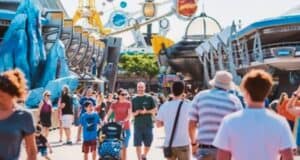 A bustling theme park with many visitors walking along a wide path. Colorful attractions, including futuristic structures and rides, are in the background, and people of all ages, including families, are enjoying their day in bright, sunny weather.