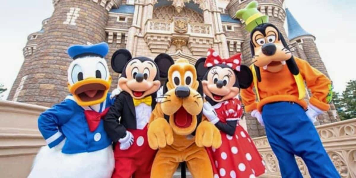 A group photo of Disney characters in front of a castle. From left to right: Donald Duck, Mickey Mouse, Pluto, Minnie Mouse, and Goofy. They are all smiling and in colorful costumes, posing cheerfully with the castle's detailed stone architecture visible in the background.