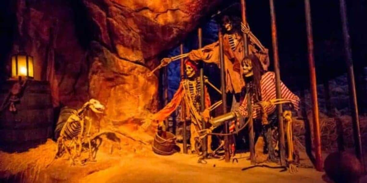 A dimly lit scene evocative of Pirates of the Caribbean features three skeletons dressed as pirates inside a cave-like cell. One skeleton holds a bone, trying to attract a dog outside the cell with keys in its mouth. The cell and surrounding area are illuminated by a lantern on a barrel.