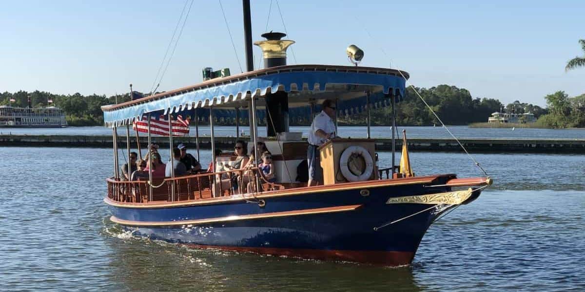 A small blue and yellow boat with an American flag at the back sails on a calm body of water on a clear day. Several passengers are seated on board, and the captain stands at the helm. In the background, beyond the shoreline with trees and buildings, lies the entrance to the Magic Kingdom Parking Lot.