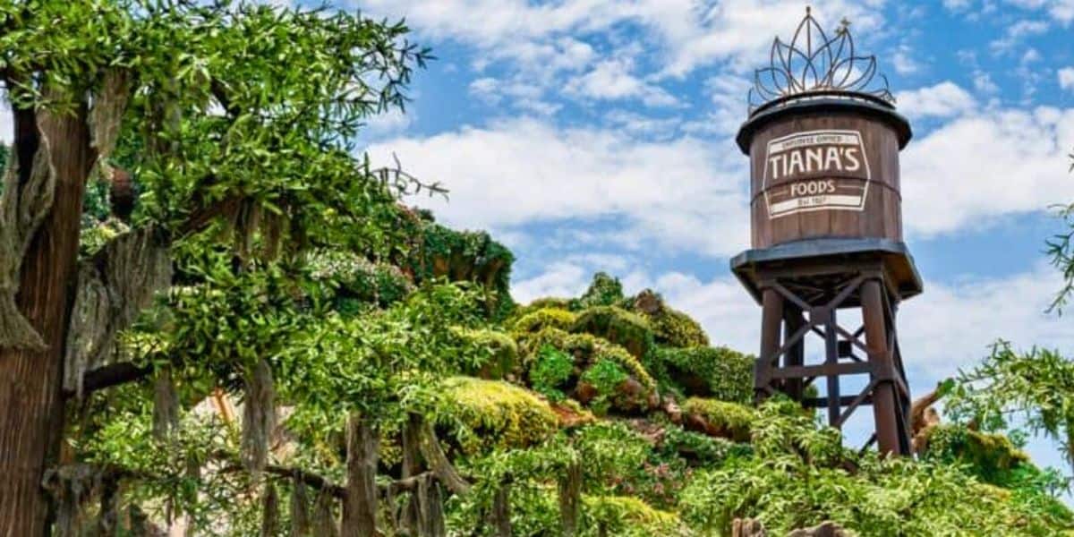 An image of a lush, green landscape with a tall water tower labeled "Tiana's Foods" prominently standing amidst the foliage. The sky is clear with a few scattered clouds, adding to the vibrant and scenic environment.