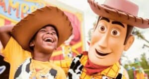 A child wearing a cowboy hat and a yellow shirt laughs joyfully next to a Disney character performer in a Woody costume from Toy Story. They are at an amusement park with colorful signs in the background, creating a lively and fun atmosphere.