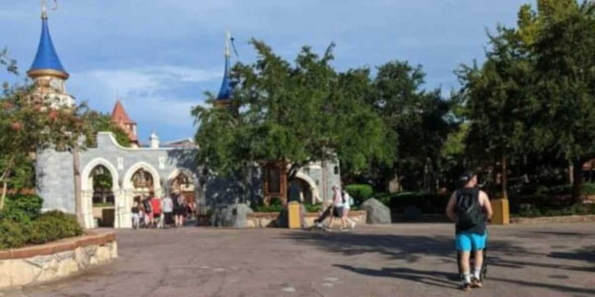 People are walking through an amusement park with castle-themed structures and colorful turrets in the background. Trees and benches are scattered throughout the scene, and the sky is clear and blue.