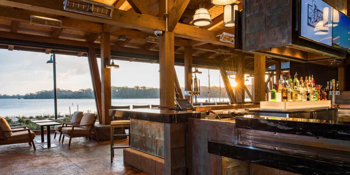 Outdoor lakeside bar with wooden beams, modern lighting, and a stocked bar counter. Comfortable seating areas overlook the tranquil lake with trees in the background. The sun is setting, casting a warm glow over the scenic setting.