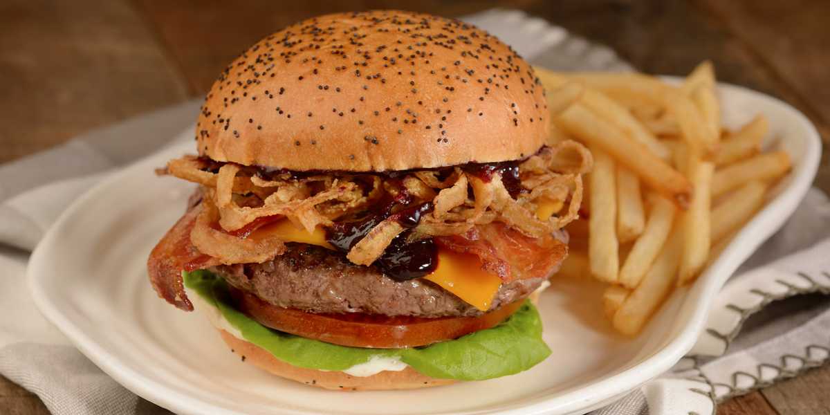 A gourmet burger with a poppy seed bun, crispy bacon, melted cheese, fried onions, and barbecue sauce, served with a side of golden French fries on a white plate.