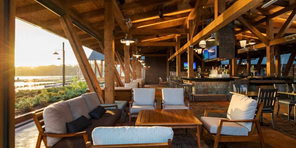 An open-air wooden pavilion with a cozy seating area featuring cushioned chairs and a coffee table, overlooking a serene lakeside view at sunset. There is a bar area in the background with high stools and television screens.
