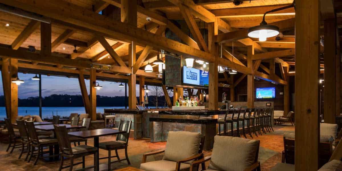 A cozy, rustic restaurant with wooden beams and warm lighting. The interior features a central bar area with stools, and seating with cushioned chairs and tables. Large windows offer a view of a serene waterfront at dusk. TVs are mounted on the walls.