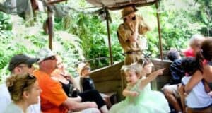 A tour guide in a safari outfit uses binoculars on a jungle-themed boat ride, engaging a group of passengers. Among them, a young girl in a fairy costume excitedly points at something, while other passengers look around and enjoy the lush green surroundings.