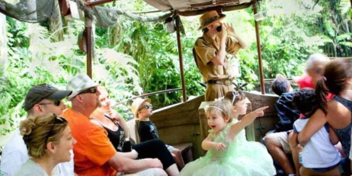 A tour guide in a safari outfit uses binoculars on a jungle-themed boat ride, engaging a group of passengers. Among them, a young girl in a fairy costume excitedly points at something, while other passengers look around and enjoy the lush green surroundings.