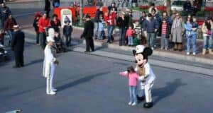 A young girl stands next to a person in a Mickey Mouse costume on a paved street, pointing to the side. A man in a white uniform with a tall feathered hat is nearby. A crowd of onlookers in casual clothing lines the sidewalk, watching the interaction.