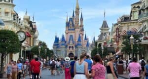 A bustling crowd walks down a wide street lined with shops and trees towards a large, ornate castle with spires and blue roofs, reminiscent of Disney World Vacations. The sky is clear, and people are enjoying their day in what appears to be a theme park.