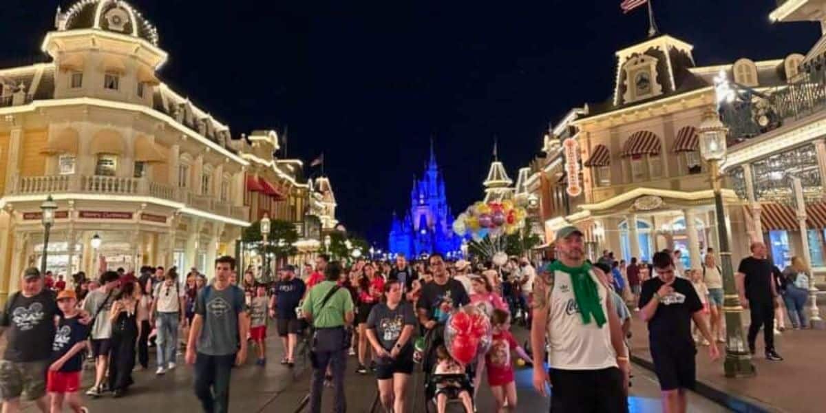 A bustling night-time scene at a theme park with visitors strolling down the main street, lit by the glow of ambient lighting, with a fairy tale castle illuminated in the background, creating a magical atmosphere.