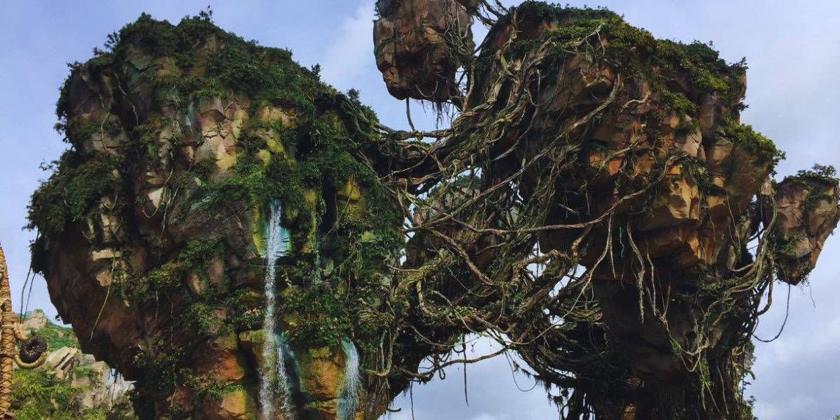 A lush, floating mountain range covered in greenery and vines, with large rocky formations connected by natural bridges. The sky is a mix of clouds and clear areas, creating a mystical atmosphere reminiscent of a fantastical landscape.