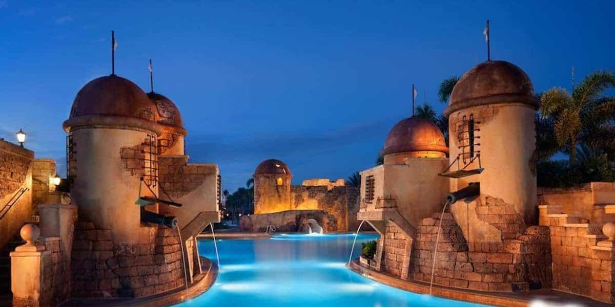 A picturesque view of a resort pool lit up at dusk, surrounded by structures resembling ancient fort towers. The serene pool water reflects the evening sky, with palm trees visible in the background, adding to the exotic ambiance reminiscent of Disney Resorts at Walt Disney World.