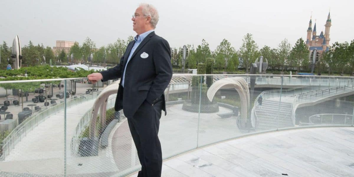 A man in a suit stands on a glass-walled terrace overlooking a park. In the background, there are buildings and trees, along with a distant castle-like structure. He appears to be speaking or addressing a group, his hands outstretched like a Disney Imagineer presenting his latest visionary project.