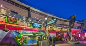 The image captures the entrance to Buzz Lightyear's Space Ranger Spin at a theme park, likely in the early evening. Illuminated by vibrant, colorful signs and lights, the area radiates a futuristic ambiance with space-themed decorations.
