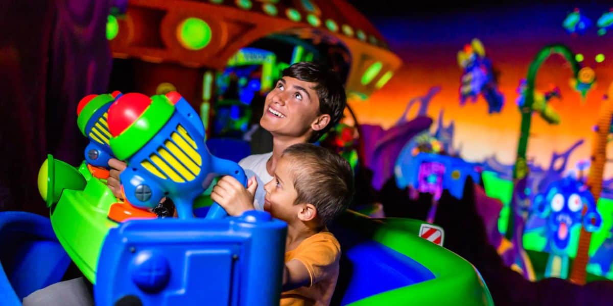 An adult and a child in a theme park ride car are joyfully interacting with colorful, futuristic toy blasters. The background is vibrant, filled with neon lights, whimsical structures, and playful aliens, reminiscent of the exhilarating Buzz Lightyear Space Ranger Spin adventure.
