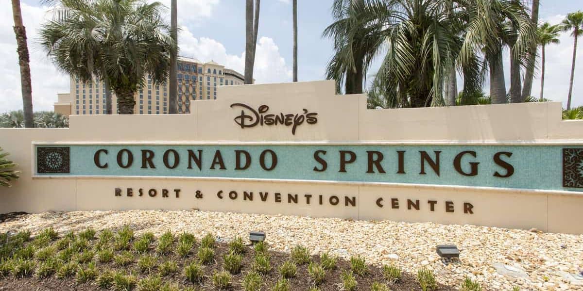 Sign for Disney's Coronado Springs Resort & Convention Center, surrounded by palm trees and greenery. The building in the background complements the sign, which features a light-colored finish and teal accents—a welcoming sight for guests of Walt Disney World.
