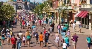 A bustling theme park scene unfolds at Disneyland Paris, with people strolling along a main street lined with shops and green lampposts. In the distance, under a clear blue sky, the iconic castle-like structure stands majestically.
